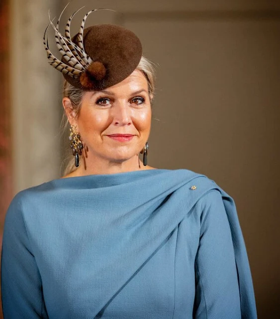 Queen Maxima wore a sky blue silk cape dress by Natan. She wore a wool camel coat. Ole Lynggaard gipsy earrings