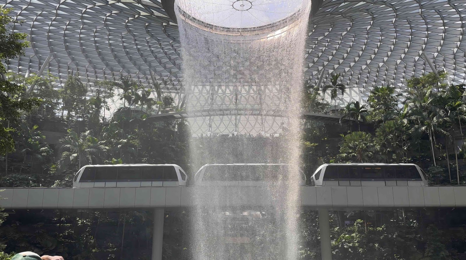 A unique view of an airport shuttle train passing directly in front of the cascading Rain Vortex waterfall inside Changi Jewel.