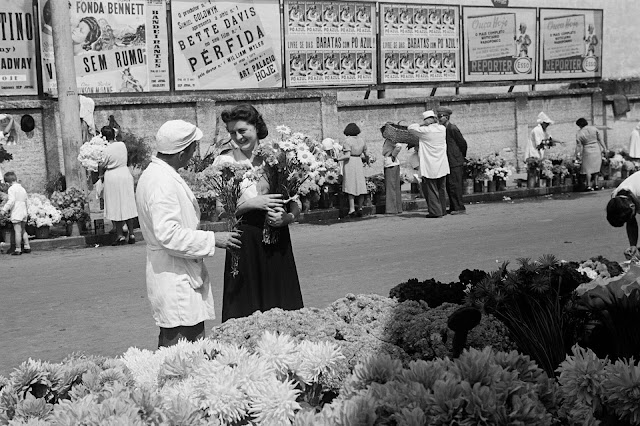 A imagem em preto e branco mostra uma feira de flores na rua. Em primeiro plano, há um vendedor de branco que carrega um pequeno buquê. Ele conversa com a modelo que sorri e segura um grande buquê em uma das mãos. Ao fundo, do outro lado da rua, vemos mais pessoas olhando as flores expostas na calçada próxima a um muro. E, acima do muro, existem diversos cartazes com propagandas expostas.