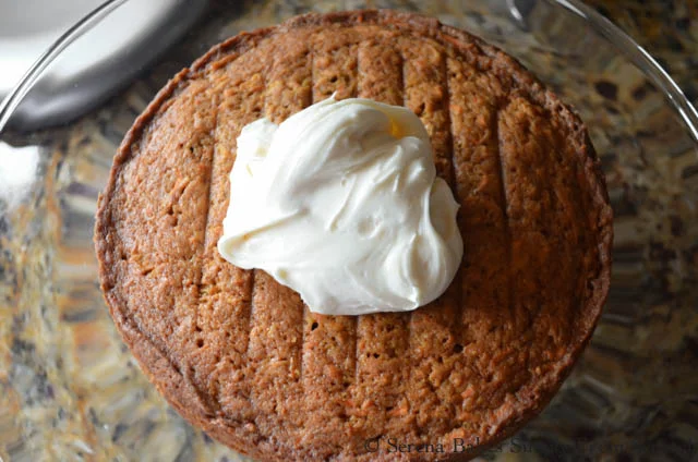 A large dollop of Cream Cheese Frosting on baked Carrot Cake with Pineapple round on a glass cake stand.