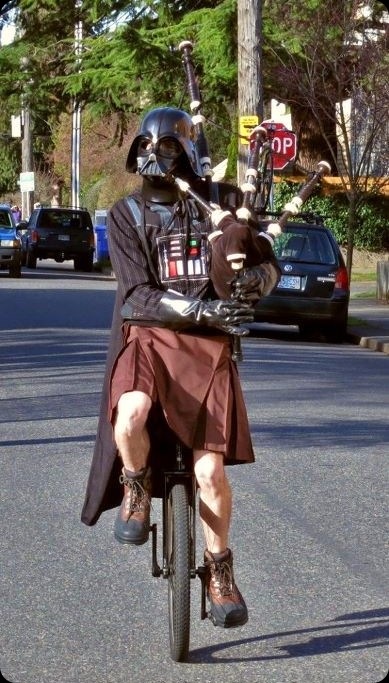 a person in a Darth Vader mask and cape, and a kilt, riding a unicycle, playing Star Wars music on a bagpipe