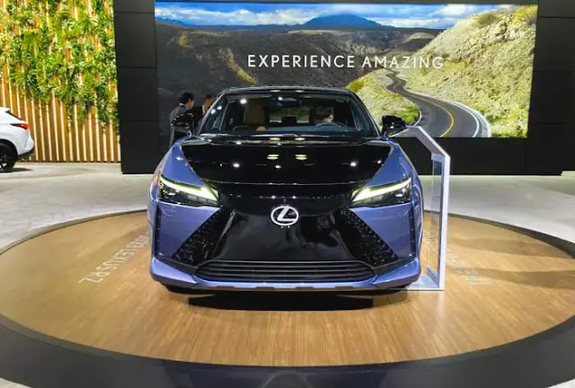 A blue Lexus RZ450e on a wood floor, with a large LED screen in the background reading "experience amazing.