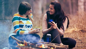 Two women sitting in a forest in front of a camp fire, chatting