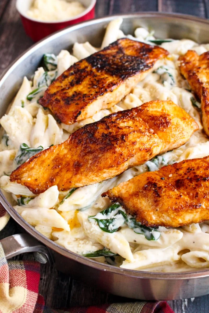 Four seared salmon fillets over cheesy spinach pasta in a stainless steel skillet on a rustic wood background.