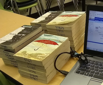 Four stacks of books consisting of two separate titles, 'Black Like Me' and 'The Red Umbrella.' Each title is grouped into two stacks each, out of the total four. The stacks of books are next to a laptop computer. The books each have a barcode in the upper-left corner, front cover. Numbers have also been written in black pen across the books' pages, bottom-edge