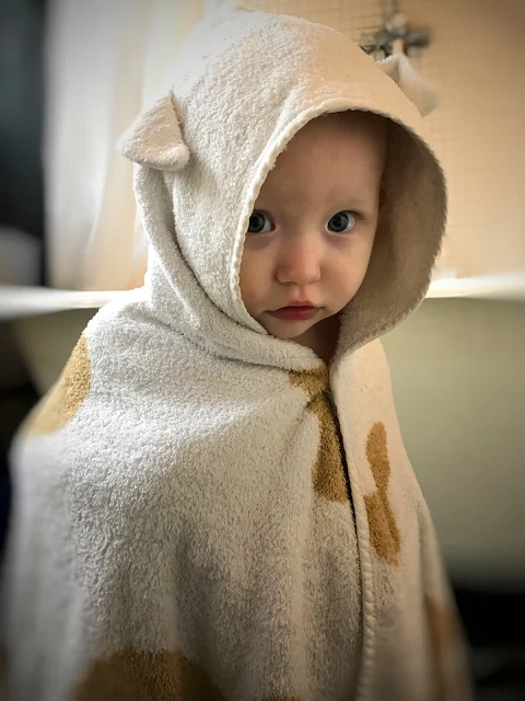 A baby in a bathroom with a roll top bath in the background. The baby is wearing a Cuddledry towel with ears and brown spots