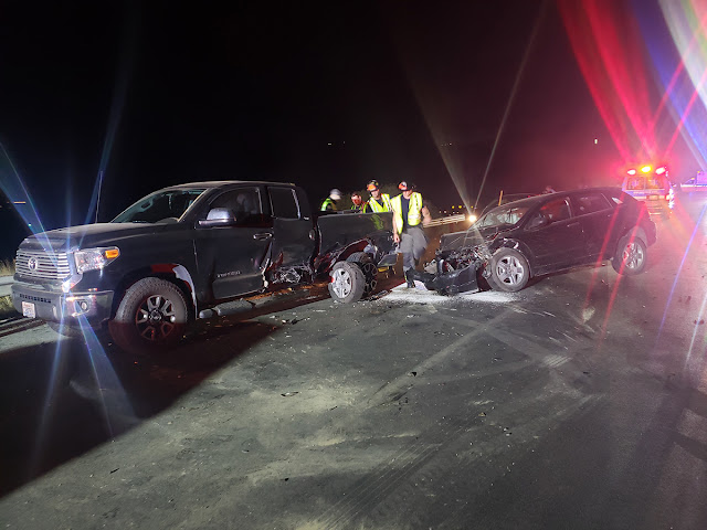 Emergency personnel wearing reflecting vests and helmets inspect the site of a crash that involved several vehicles including a black pickup truck and a black sedan. In the upper right-hand corner, the lights from emergency vehicles light up the night sky.