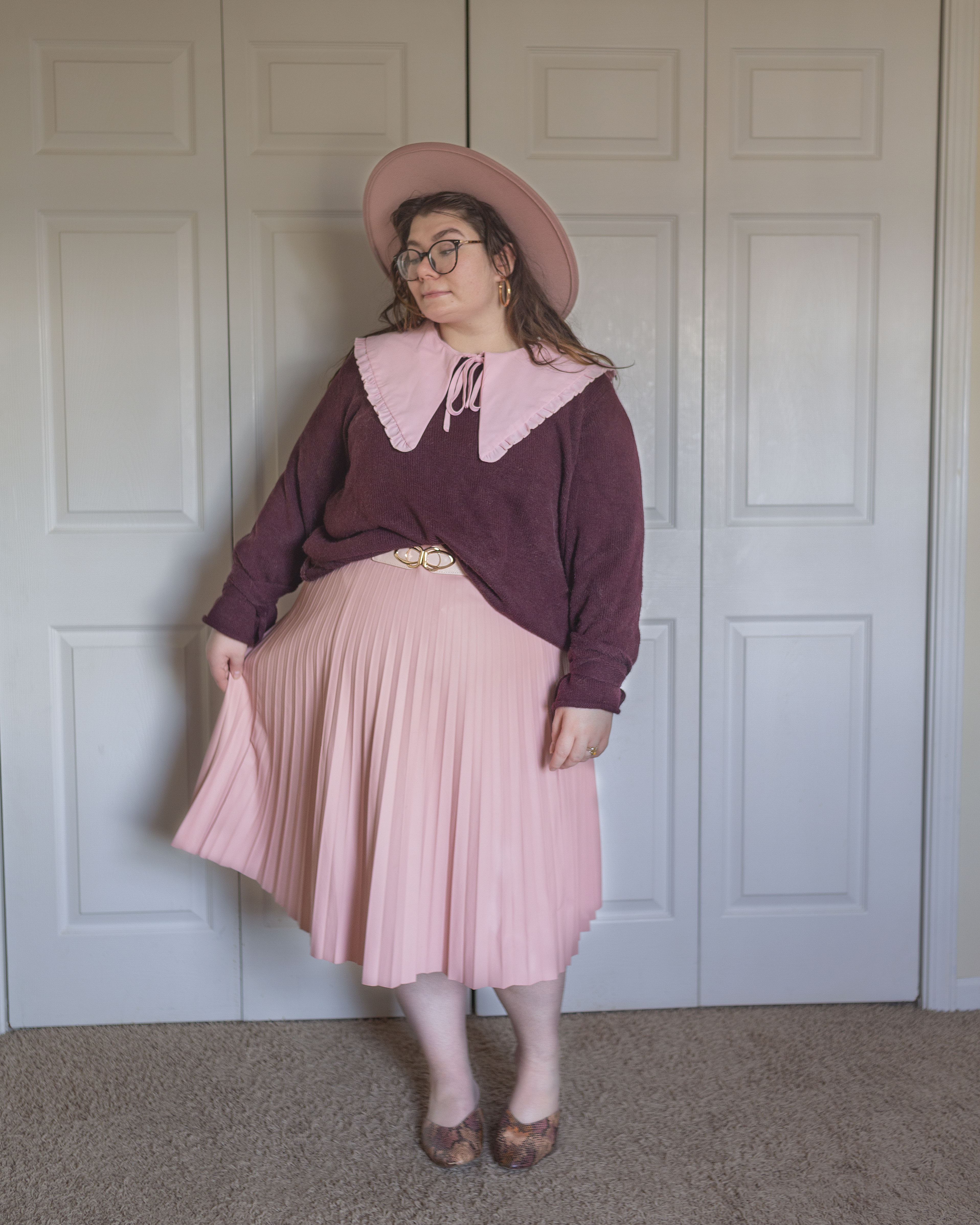 An outfit consisting of a pastel pink wide brim fedora hat, a pastel pink oversized peter pan collared blouse with ruffles at the hem of the collar under a plum colored knit sweater that ties in bows around the wrist half tucked into a pastel pink accordion pleated faux leather midi skirt and pink, brown, and black faux snake skin mules.