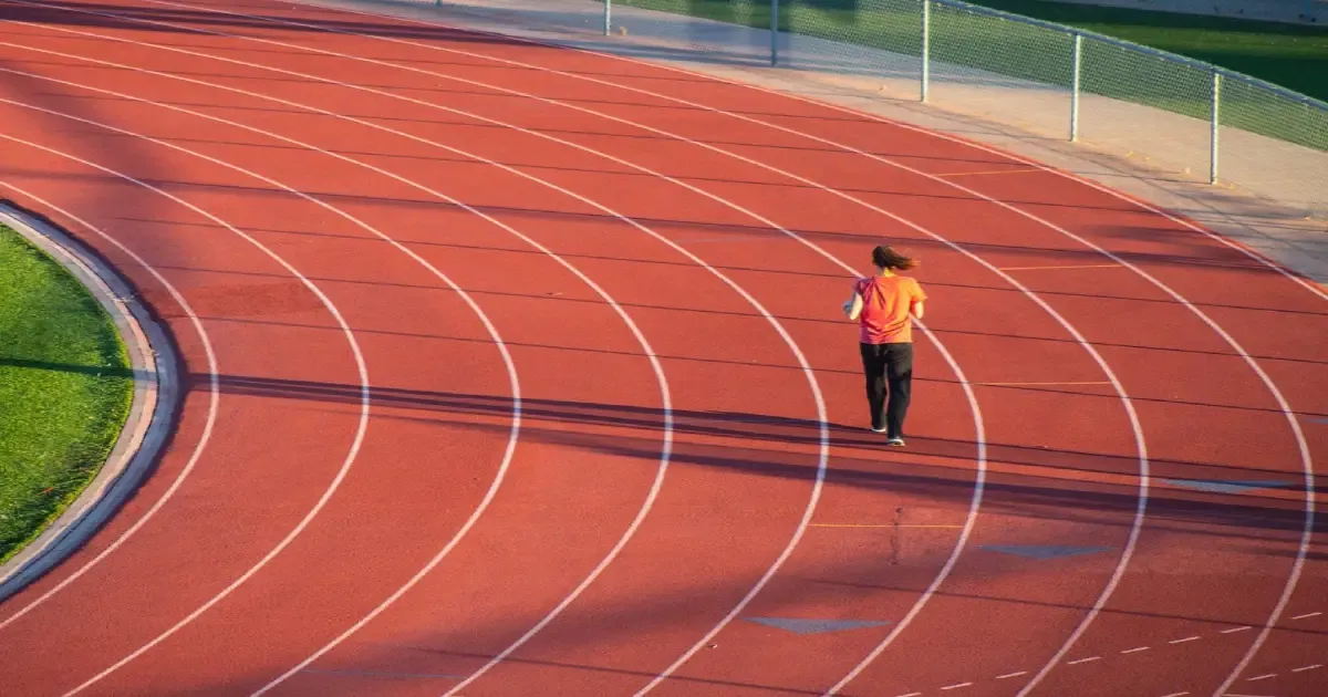 Person jogging alone on a sunny outdoor athletic track beside a green field.