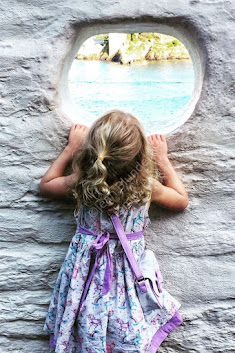 A small blonde girl in a lilac and white patterned dress peeps through a hole in a whitewashed wall and sees the sea and rocks beyond