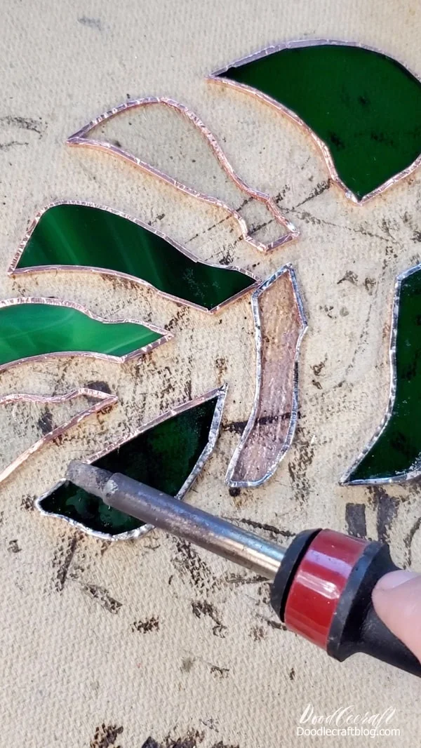 Okay, set your stained glass monstera leaf pieces on your fire resistant surface.   Open up the flux and pour some of the solution into the cap.    Dip the flux brush into the cap and brush it on all the pieces, right on the copper foil.   Then touch the soldering iron to the solder briefly and then rub the soldering iron on the edge of the glass pieces.   The solder goes pretty far and needs just a light coating over all the copper foil--this will make it easier for all those holes and curves in the leaf.