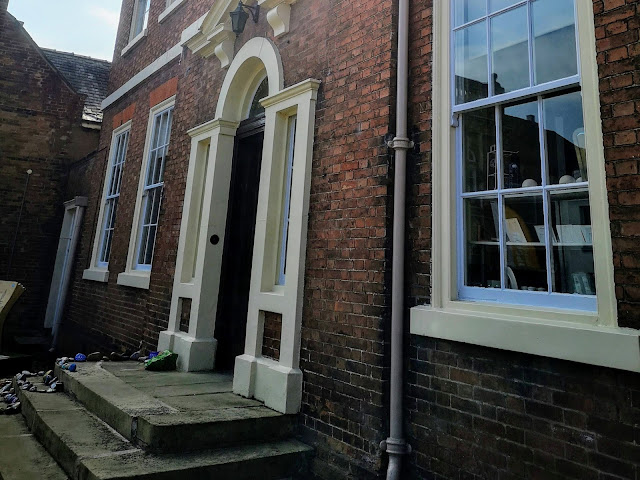 The front of Amcott House, home of Bassetlaw Museum. The image shows a tall listed building made of red brick with large leaded windows that are painted white.There is a decorative archway framing a large heavy wooden black door. There are steps up to the door. Alongside the steps are some stones that have been painted throughout lockdown and placed there by the public.