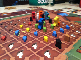The board in the middle of a game. The focus of this photo is the central area of the board, a plot of farmland represented by several brown squares in a grid. Each square has a cutout in the shape of a chili pepper. Some of these have wooden pepper-shaped tokens in various colours in them. There are five farmer-shaped meeples in the board, standing between two squares on the board.