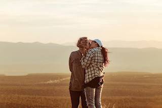 Two members of the public kissing affectionately in the middle of a light coloured field with flowers around them and a blue sky on a light background. 