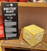 A close up photo of a square gold cube bath bomb with a question mark in the middle on a light brown shelf with a black rectangular card with question block lush x super Mario bros movie in white font on a bright background