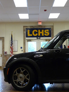 The front of car sits on the inside of a building with a tiled floor. Behind it is doors to the outside, framed in marble, and an american flag. A banner of the doors say "LCHS."