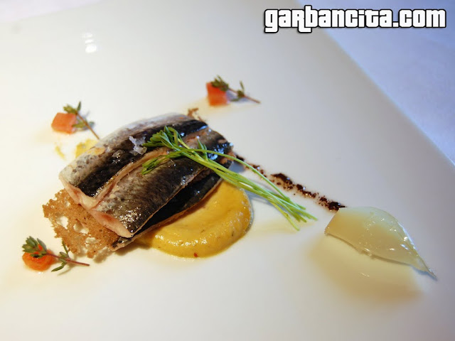 Sardinas cocinadas en humo de haya, sobre trazo de encurtidos, anchoas, olivas negras, germinados de lenteja y cebolleta asada