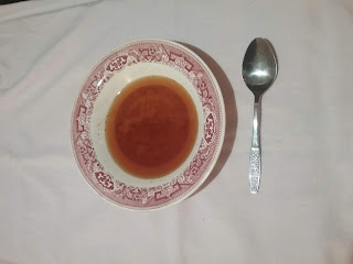 A shallow soup bowl of pink transferware, containg a light orange-brown broth. A silver-colored spoon is next to the bowl.