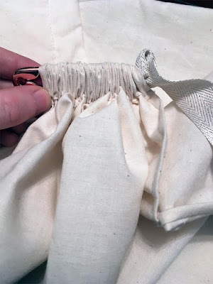 A length of white muslin scrunched tightly onto a rose-gold-colored safety pin, with the head of the pin just emerging from one end and a length of white and gold herringbone tape at the other.