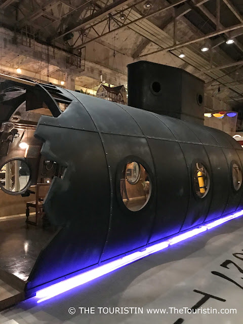 A piece of a black submarine with portholes, illuminated by a blue tube lamp, parked in a giant factory hall with concrete walls.