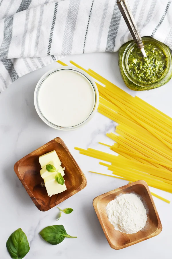 An assortment of ingredients for preparing creamy pesto pasta displayed on a luxurious marble countertop. The ingredients include fragrant pesto sauce, creamy dairy, pasta, butter, flour, and a bunch of fresh basil leaves.