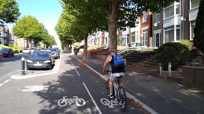 A steep hill. The two-way cycle track is on the fright hand side of the street with car parking left and a footway with houses beyond to the right.