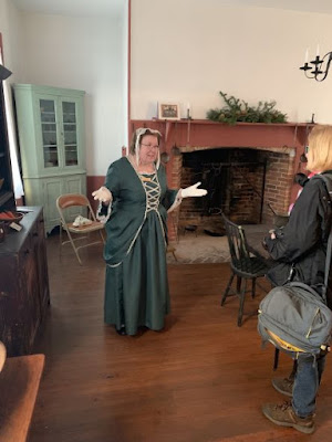 White woman with short dark hair and glasses stands in front of a large fireplace wearing a dark green colonial-style dress with white edging, white gloves and a white cap. She is talking to a visitor wearing a dark gray jacket.