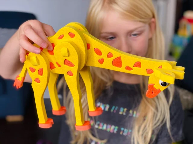 A young girl holding a yellow and orange foam giraffe having assembled the puzzle from lots of different flat pieces. The giraffes neck is pointing down