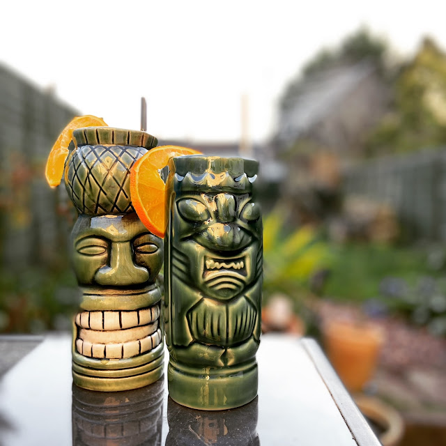 Two green tiki mugs standing on a glass table, with a garden stretching out in the background.