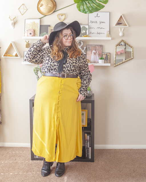 An outfit consisting of a black wide brim hat, an animal print blouse with an attached black ribbon tied into a bow, tucked into a yellow button down skirt and black Chelsea boots.