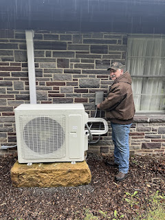 A man wearing jeans, work boots, a brown leather jacket, and a camo cap stands next to a stone house connecting an HVAC system.