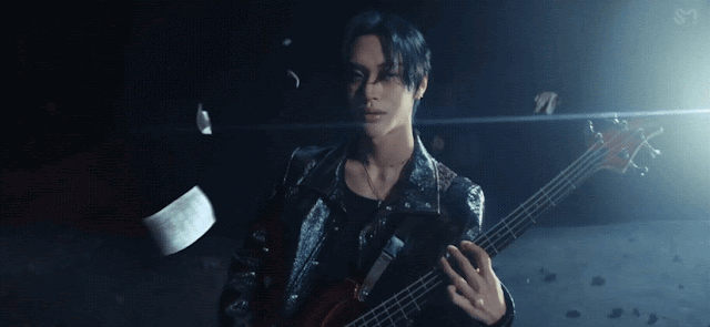 Gif from the music video "Boom Boom Bass" by Kpop boy group Riize where Wonbin is in space playing the bass and smirks at the camera. There are cups, papers and basses flying around him