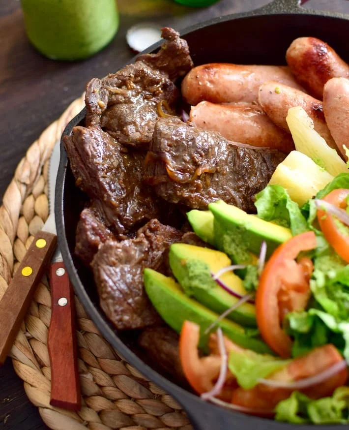 La parrilla venezolana es sinónimo de convite, celebración familiar y con amigos. Es una comida abundante. Carne y chorizos, ensalada criolla verde con aguacate, y yuca, hecha y servida en una sartén de hierro