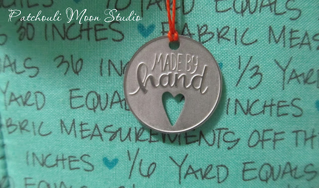 closeup of metal tag that says "made by hand" and depicts a cut out heart