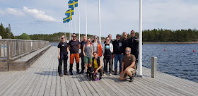 12 ihmistä ja 1 koira ryhmäkuvassa laiturilla, taustalla liehuu Ruotsin lippuja. 