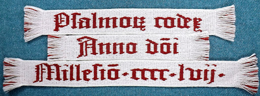 A photo of a piece of blue fabric on which sits a white tablet woven band with red lettering that reads "Psalmorum codex Anno domini Millesimo cccc lvij" or "Book of psalms 1457"