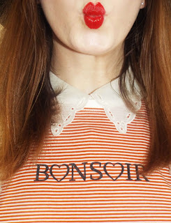 Framed shot of Ellie's pouting red lip, white vintage detailed collar and striped orange and white T-shirt layered over, that reads "Bonsoir" in black font, with the letters o in heart shapes..