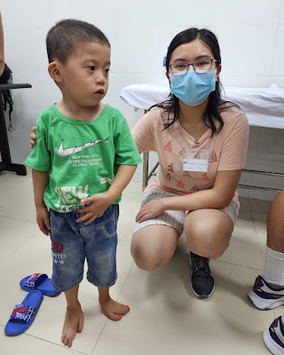 A small boy wearing a green shirt standing with a woman in a surgical mask while waiting to have his foot evaluated for surgery as part of an International Extremity Project medical mission.
