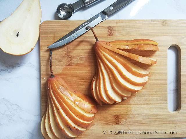 Sliced pear fans on a cutting board with knife, pear half and melon baller