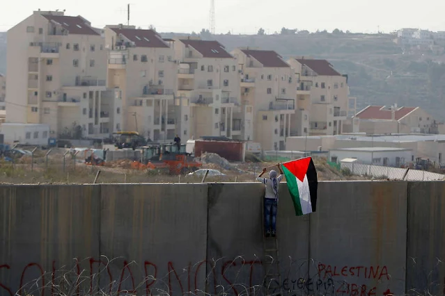 رجل يضع العلم الفلسطيني على الجدار العازل الإسرائيلي المثير للجدل، وتظهر المستوطنات الإسرائيلية في الخلفية