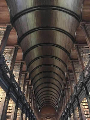 A shot straight down the center of a two-storey library with tall shelves of old, brown and taupe books on either side, and a section of molding separating the two levels of shelves on either side. The dark wood center ceiling is a half-round, spanned by dark arches that spring from the tops of the upper bookshelves. Between each upper set of shelves, the ceiling is lighter, though still arched and paneled in wood.