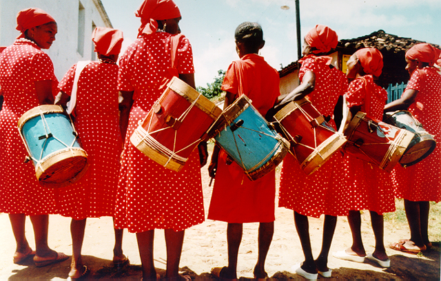 As mulheres estão de costas, todas com um mesmo vestido. Elas usam lenços na cabeça e carregam consigo uma instrumento de percussão, tambor.