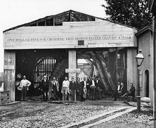 A group of men stand on a cobblestone street at the entrance of the covered bridge. “One dollar fine for crossing this bridge faster than on a walk,” is written across the top in all capital letters.