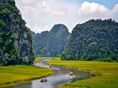 The Tam Coc (“three caves”) portion is a three-hour excursion by small boat along the Ngô Đồng river,  beginning at the village of Van Lam and proceeding through a scenic landscape dominated by rice fields and karst towers. The route includes floating through three natural caves (Hang Ca, Hang Hai, and Hang Ba), the largest of which is 125m long with its ceiling about 2m high above the water. The boats are typically rowed by one or two local women who also sell embroidered goods.