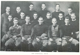 A black and white photograph of seventeen seated men in dark clothes. One in the front holds a football with "1915" painted on it. The photograph is captioned "1915 Football team."