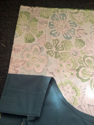 A close up of the bib of a colonial-blue cooking apron, folded down to reveal the marked pattern. Faint pen lines descibe a square neckline and lightly curved outer edge against the faintly patterned back of the green, turquoise, and grey tropical floral fabric.