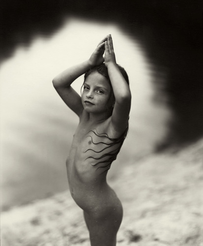 A fotografia em preto e branco mostra uma menina com os braços acima da cabeça, ela está nua e seus longos cabelos formam um desenho na lateral esquerda do seu corpo. Ao fundo podemos ver um lago e sua orla de areia, local onde a menina está de pé.