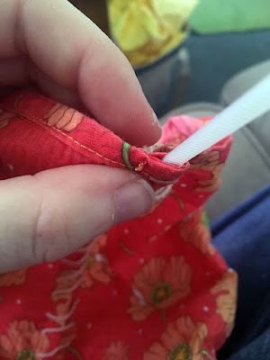 A close-up of a white hand holding the bound edge of a piece of coral fabric with scattered orange poppy motifs. At the right edge of the fabric, the folded-over seam allowance is open, and a translucent zip tie is being inserted. In the background, a canvas bag full of yellow fabrics is blurrily visible.