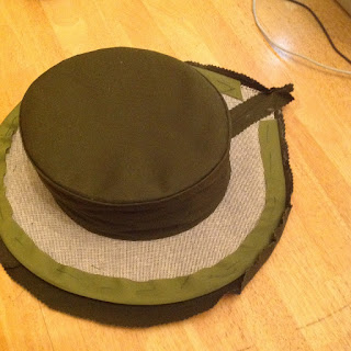 Image is of a hat about half-finished. The top part is covered in dark green fabric, but the brim has a layer of white buckram and light green fabric exposed.