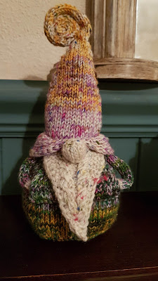Knitted Gnome on a shelf. She has her hands in a pocket on the her front, hidden by her beard.
